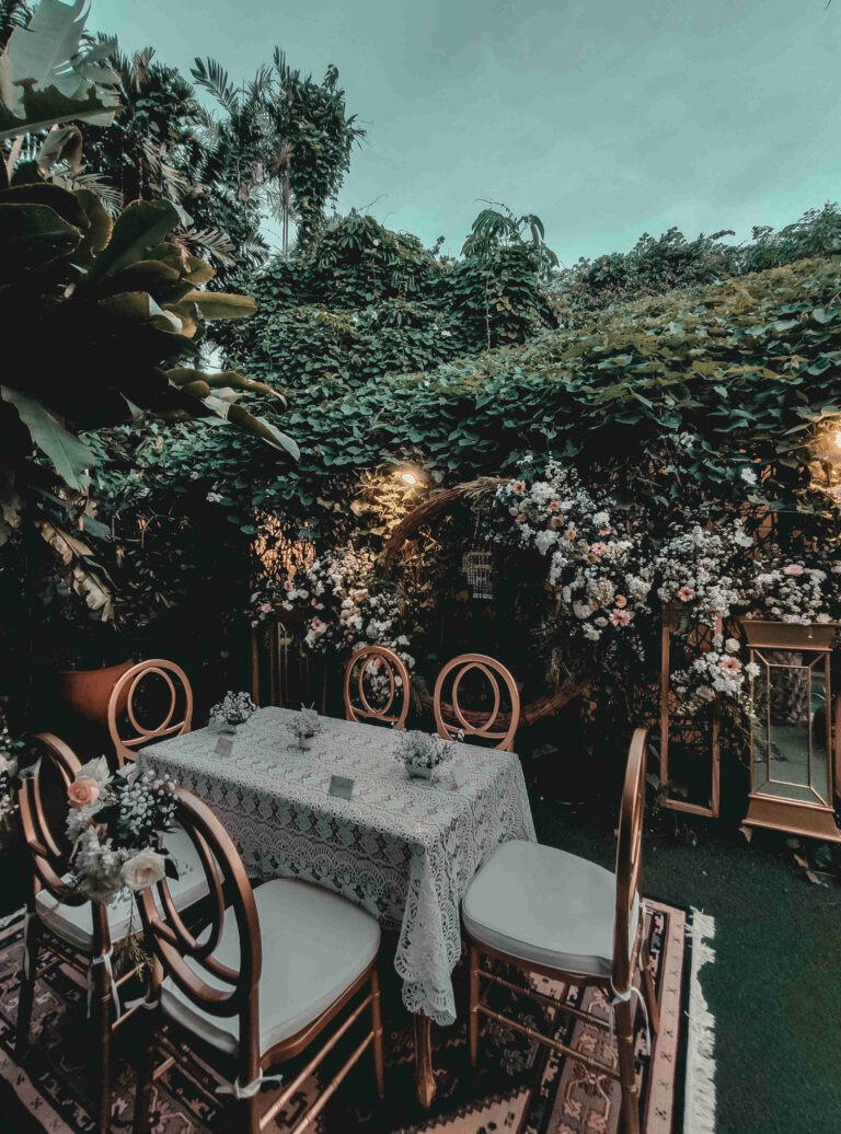 An elegantly set table with a lace tablecloth and floral centerpieces is situated in a lush garden surrounded by dense green foliage and blooming flowers. The table is adorned with five ornate chairs, and soft lighting enhances the serene ambiance. The backdrop features a decorative arch covered in flowers, adding to the romantic and enchanting atmosphere of the outdoor setting. This scene exemplifies sophisticated birthday ideas in Charlotte NC, for adults, offering beautiful and serene birthday party ideas in Charlotte NC for adults.