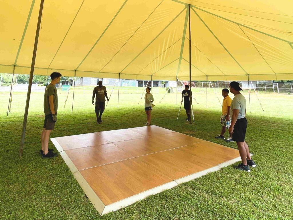 Group of six people near a wooden dance floor setup outdoors under a tent on a grass field - Dance Floor Rentals Kannapolis NC.