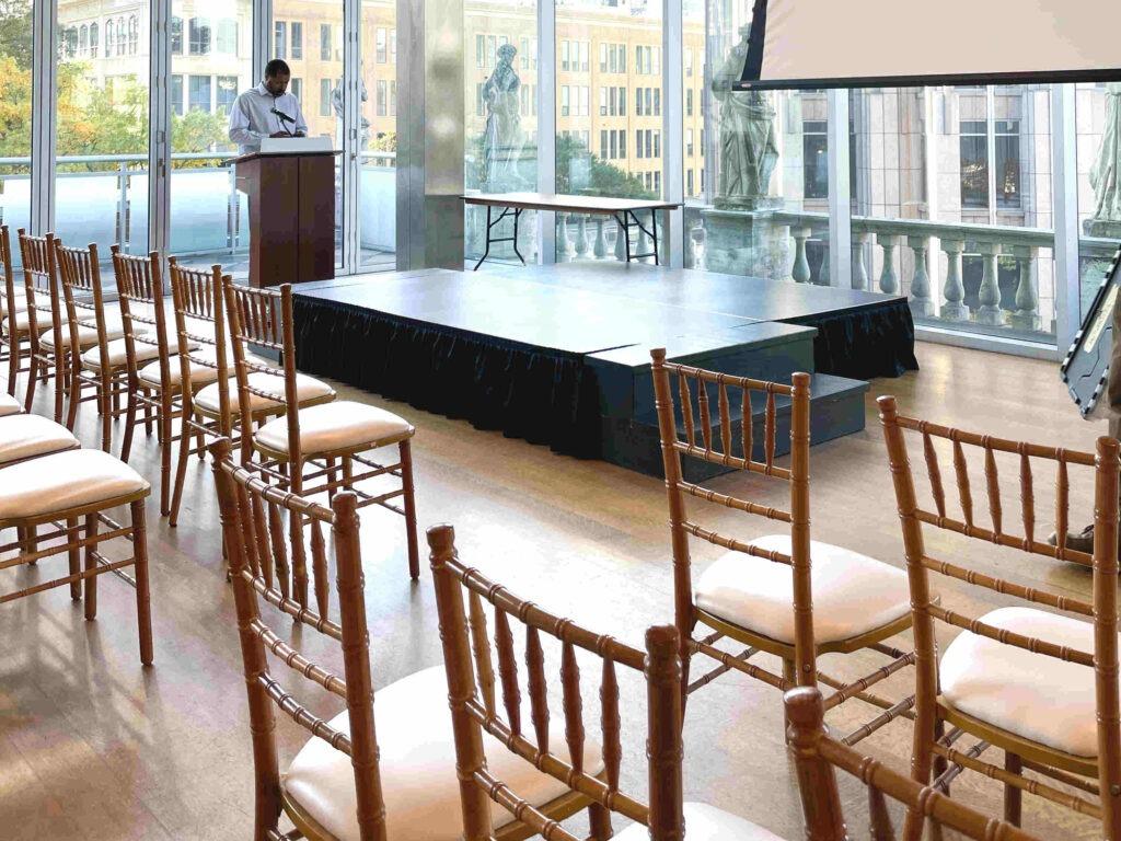 Wide windowed conference room with wooden chairs, a black front stage, and a man preparing for a speech, exemplifying professional Staging Rentals Charlotte NC.